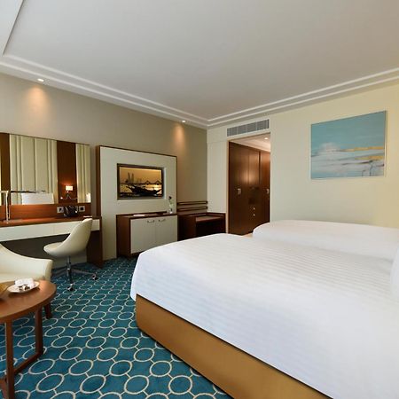 Grand Swiss-Belhotel Waterfront Seef Sanabis Exterior photo The photo shows a well-decorated hotel room featuring two beds with white linens. There's a comfortable seating area with a small round table that holds a bowl of fruit. A stylish chair is positioned next to the table. The room has a modern design wi
