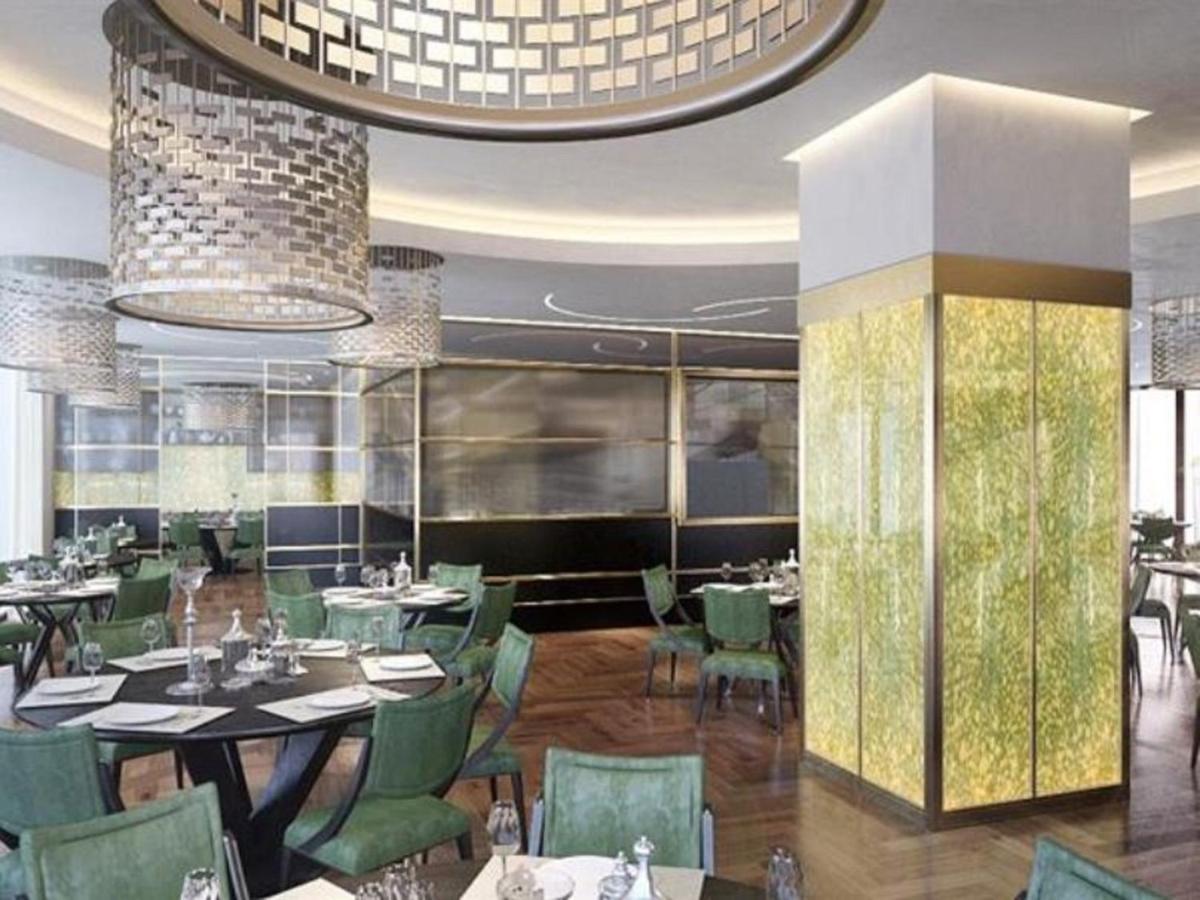 Grand Swiss-Belhotel Waterfront Seef Sanabis Exterior photo The photo shows the interior of a modern restaurant. It features a sophisticated design with stylish, green upholstered chairs arranged around dark tables. The dining area is illuminated by large, elegant light fixtures hanging from the ceiling. Ther