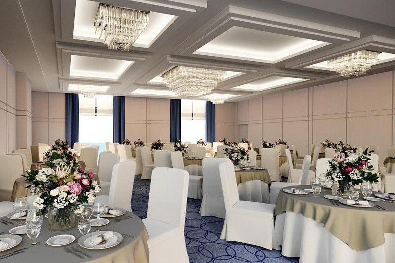 Grand Swiss-Belhotel Waterfront Seef Sanabis Exterior photo The photo shows an elegant banquet hall or event space. There are several round tables covered with light-colored tablecloths, each adorned with floral centerpieces featuring an array of flowers. The chairs are dressed in white covers. The room is we