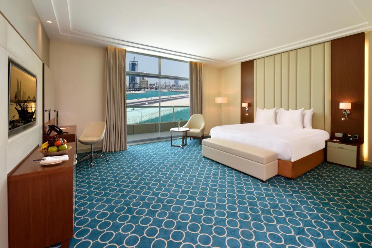 Grand Swiss-Belhotel Waterfront Seef Sanabis Exterior photo The image shows a well-lit hotel room featuring modern decor. There is a large bed with white bedding, a bedside table with lamps on either side, and a stylish area rug with a circular pattern in shades of blue and white. A small seating area include