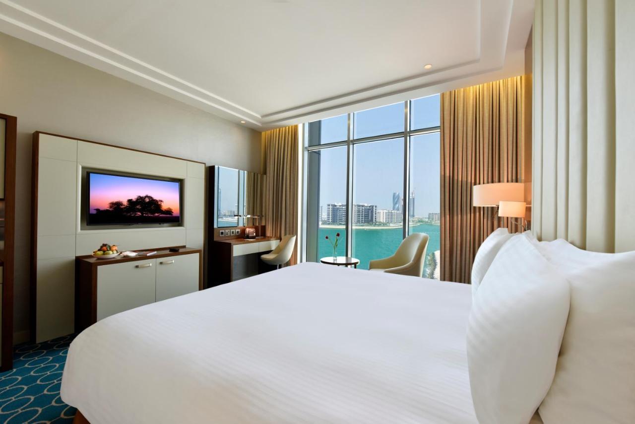 Grand Swiss-Belhotel Waterfront Seef Sanabis Exterior photo The image shows a modern hotel room featuring a large, neatly made bed with white bedding. There is a wall-mounted television opposite the bed, and a small desk with a chair is situated near large windows that appear to offer a view of the outside. T