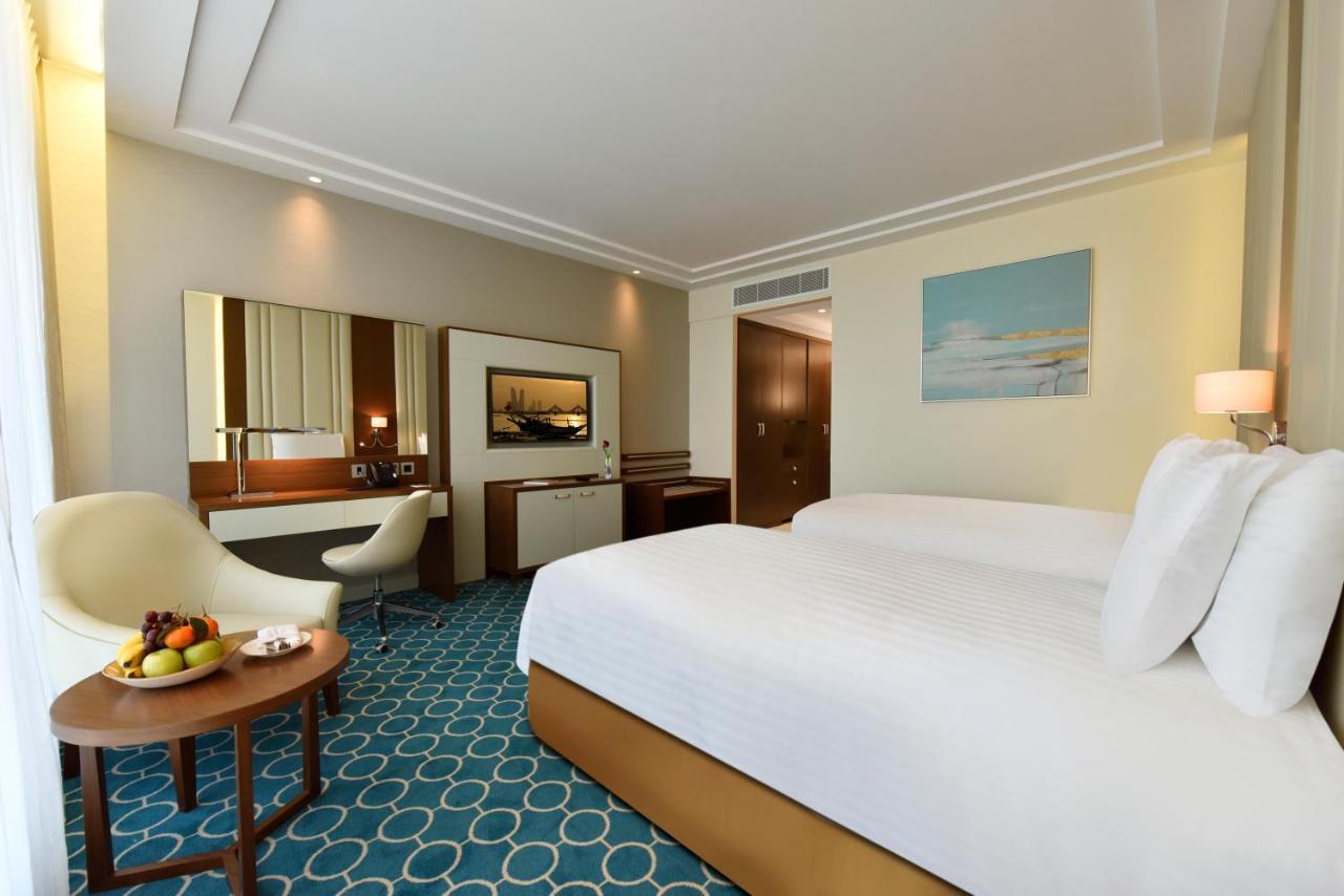 Grand Swiss-Belhotel Waterfront Seef Sanabis Exterior photo The photo shows a well-decorated hotel room featuring two beds with white linens. There's a comfortable seating area with a small round table that holds a bowl of fruit. A stylish chair is positioned next to the table. The room has a modern design wi