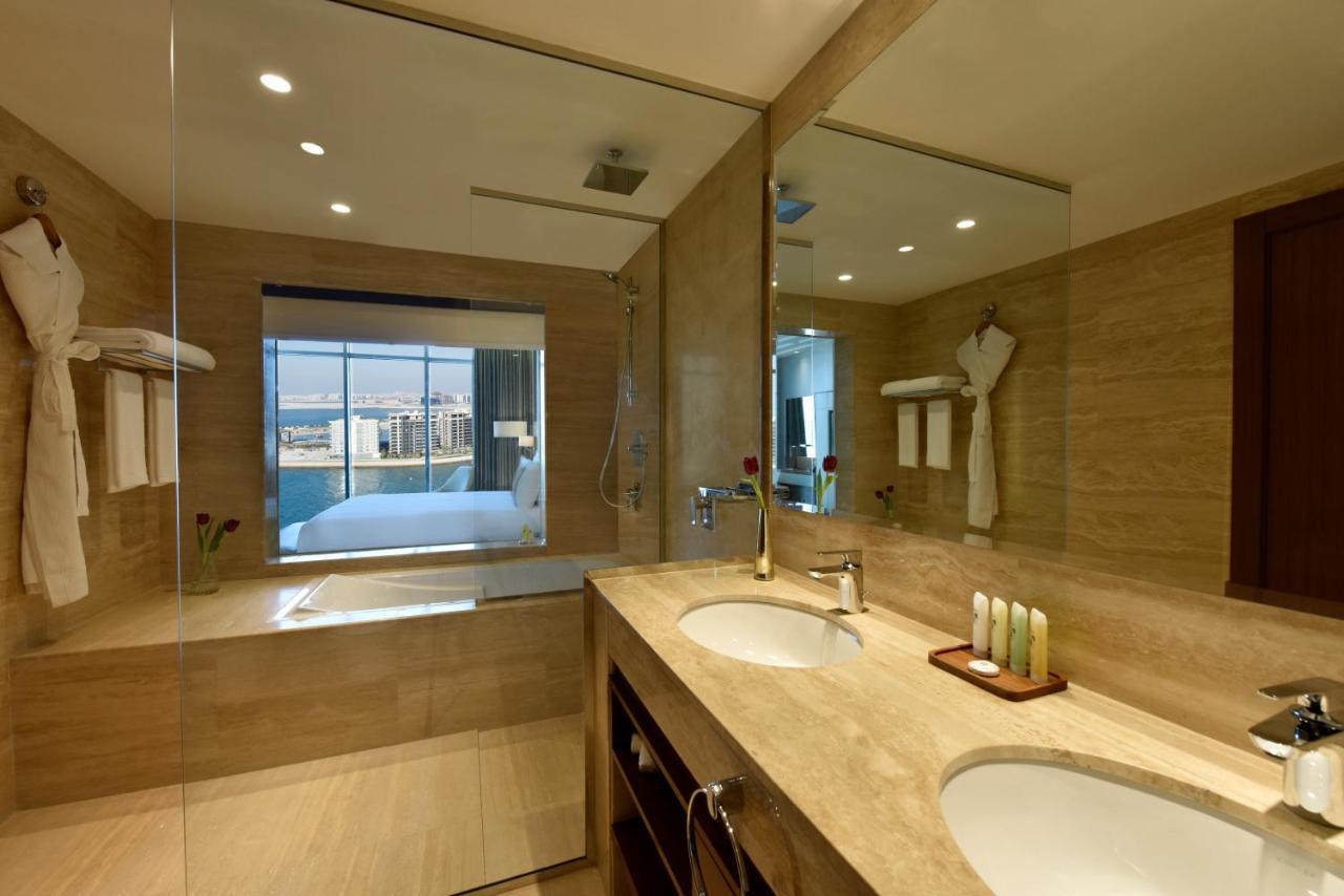 Grand Swiss-Belhotel Waterfront Seef Sanabis Exterior photo The photo depicts a modern bathroom with a sleek design. It features a large mirror above a double sink with a wooden cabinet below. The walls are adorned with light-colored tiles, and there is a glass shower enclosure. On one side of the room, there