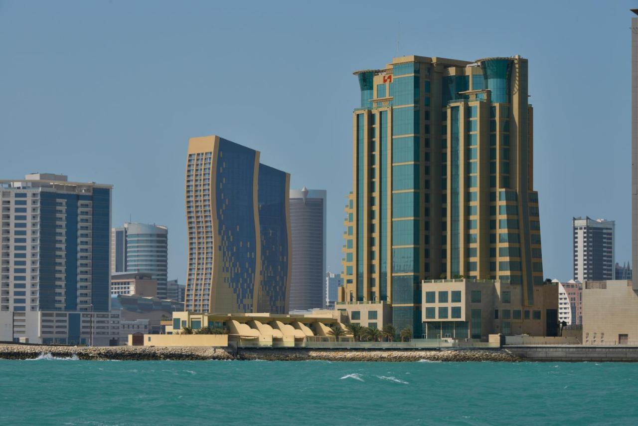 Grand Swiss-Belhotel Waterfront Seef Sanabis Exterior photo The photo showcases a coastal city skyline featuring several modern buildings. Prominent in the foreground is a tall structure with a distinctive design, characterized by a mix of green, gold, and glass. Next to it is another building with a more cur