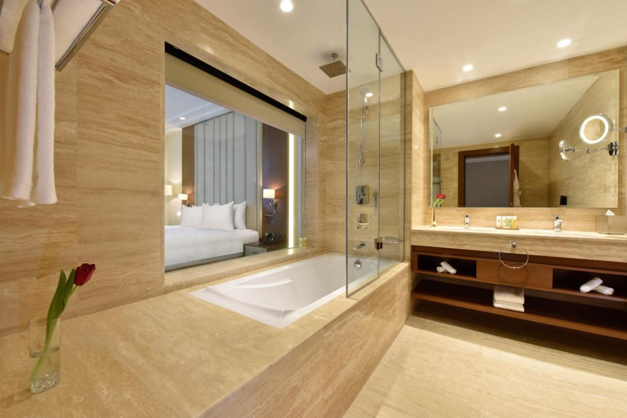 Grand Swiss-Belhotel Waterfront Seef Sanabis Exterior photo The photo shows a modern bathroom with a sleek design. It features a bathtub, which is partially enclosed by glass. The walls are made of light-colored stone, giving a clean and elegant look. There is a large mirror above the vanity, which has some l