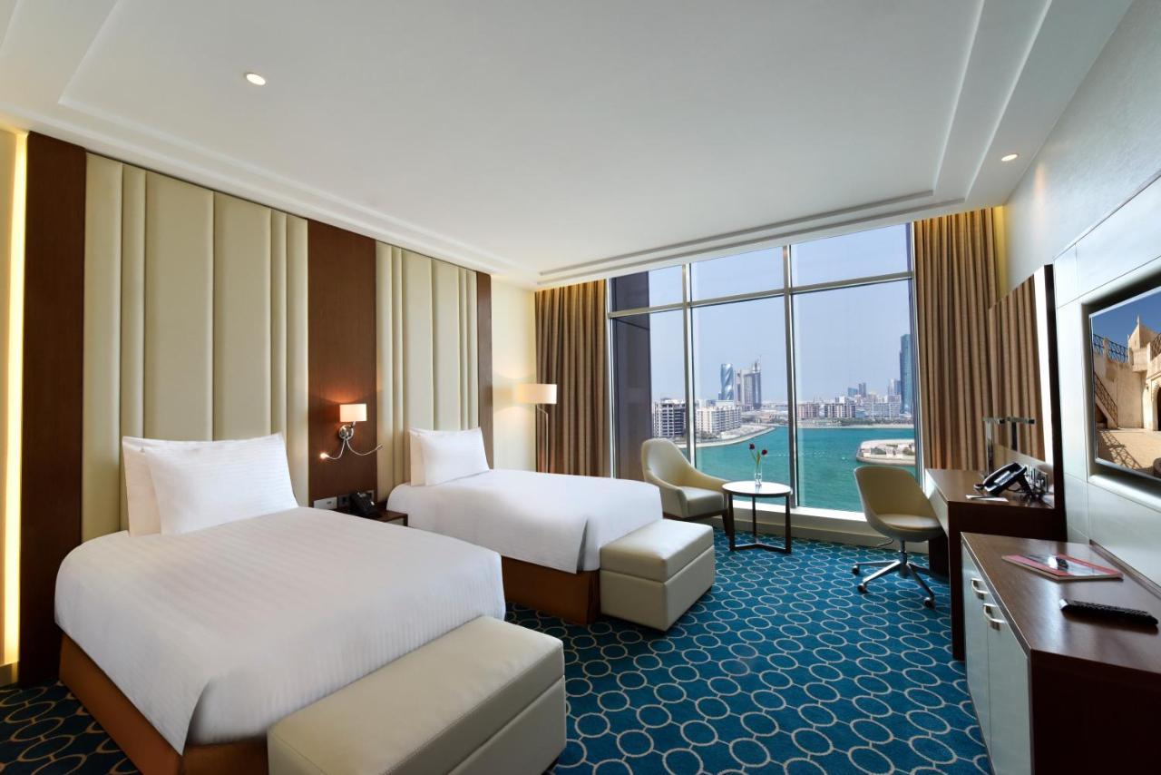 Grand Swiss-Belhotel Waterfront Seef Sanabis Exterior photo The photo shows a hotel room featuring two twin beds with white bedding. The interior design includes soft, neutral tones on the walls, accented by a textured panel. There are large windows that offer a view of a body of water and city skyline in the