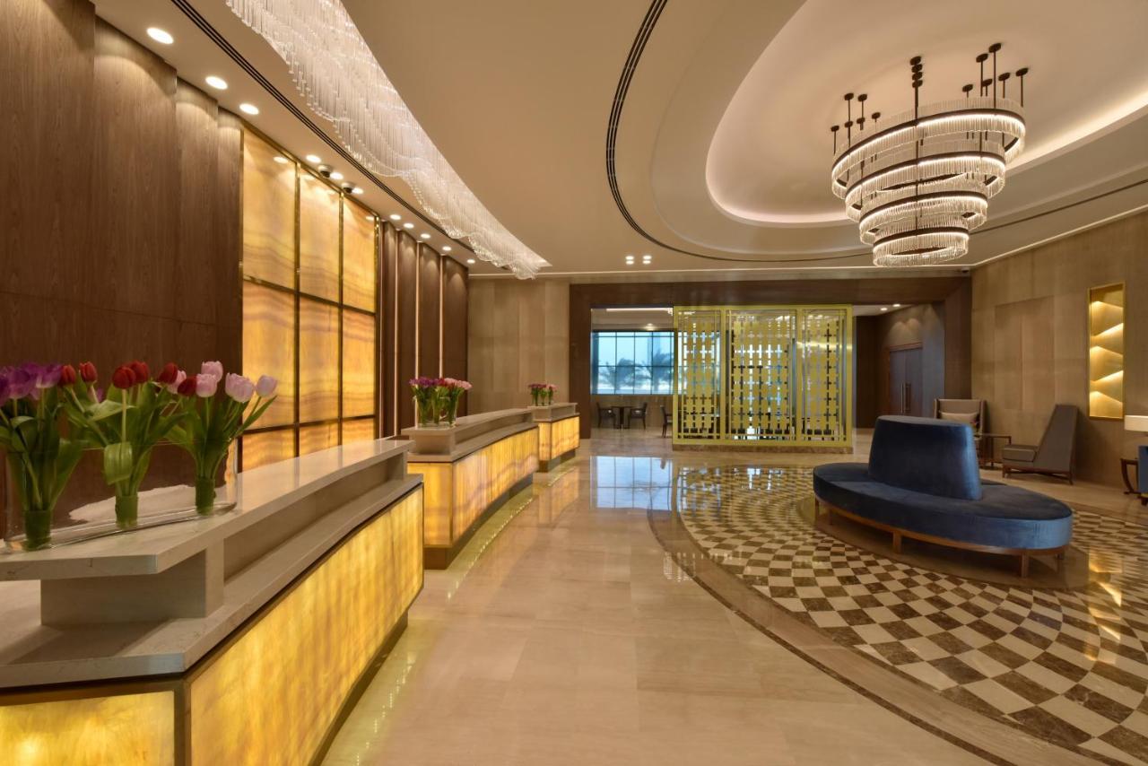 Grand Swiss-Belhotel Waterfront Seef Sanabis Exterior photo The photo shows an elegant hotel or lobby area. There is a reception desk with a warm yellow glow, flanked by potted flowers, possibly tulips. The interior features a modern design with a sophisticated chandelier hanging from the ceiling. To the side