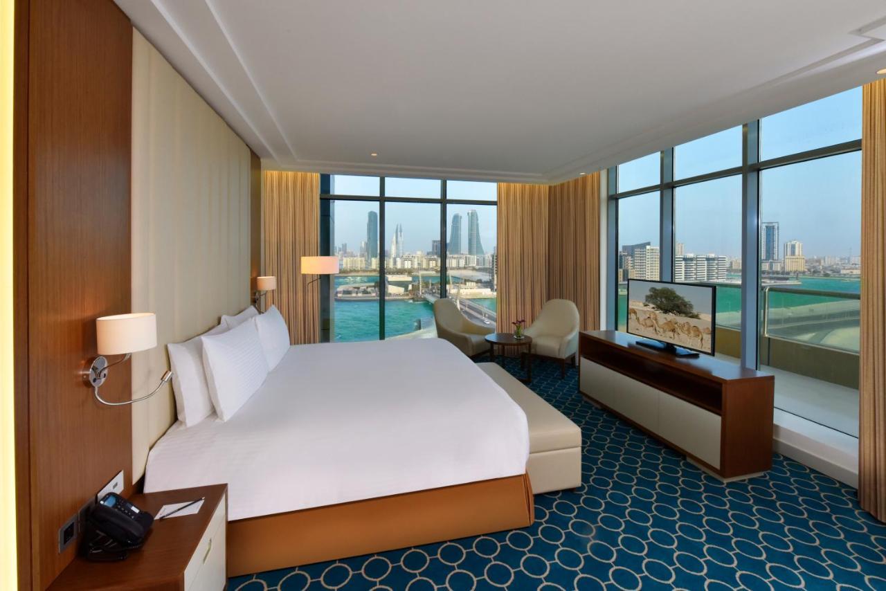 Grand Swiss-Belhotel Waterfront Seef Sanabis Exterior photo The photo shows a modern hotel room featuring a large bed with white linens positioned against a wall. There are large windows offering a view of the city and water beyond, allowing natural light to fill the space. The room includes a small seating a