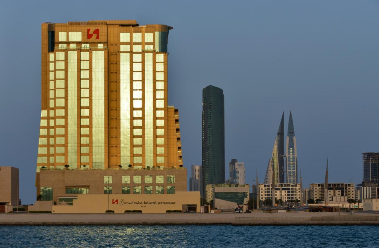 Grand Swiss-Belhotel Waterfront Seef Sanabis Exterior photo The image shows a waterfront cityscape featuring several buildings. In the foreground, there is a tall building with a golden exterior, reflecting sunlight. This building appears to be modern and is likely used for commercial purposes. Behind it, the