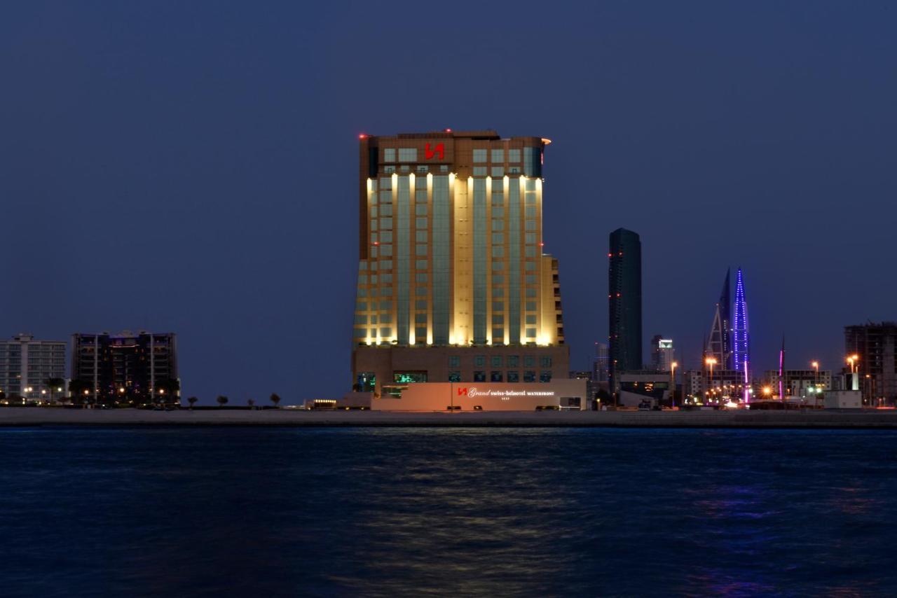 Grand Swiss-Belhotel Waterfront Seef Sanabis Exterior photo The photo shows a tall building illuminated at night, with a blend of warm yellow and cool white lights. The structure has a modern architectural design and appears to be a hotel or high-rise establishment. It is situated near a body of water, likely