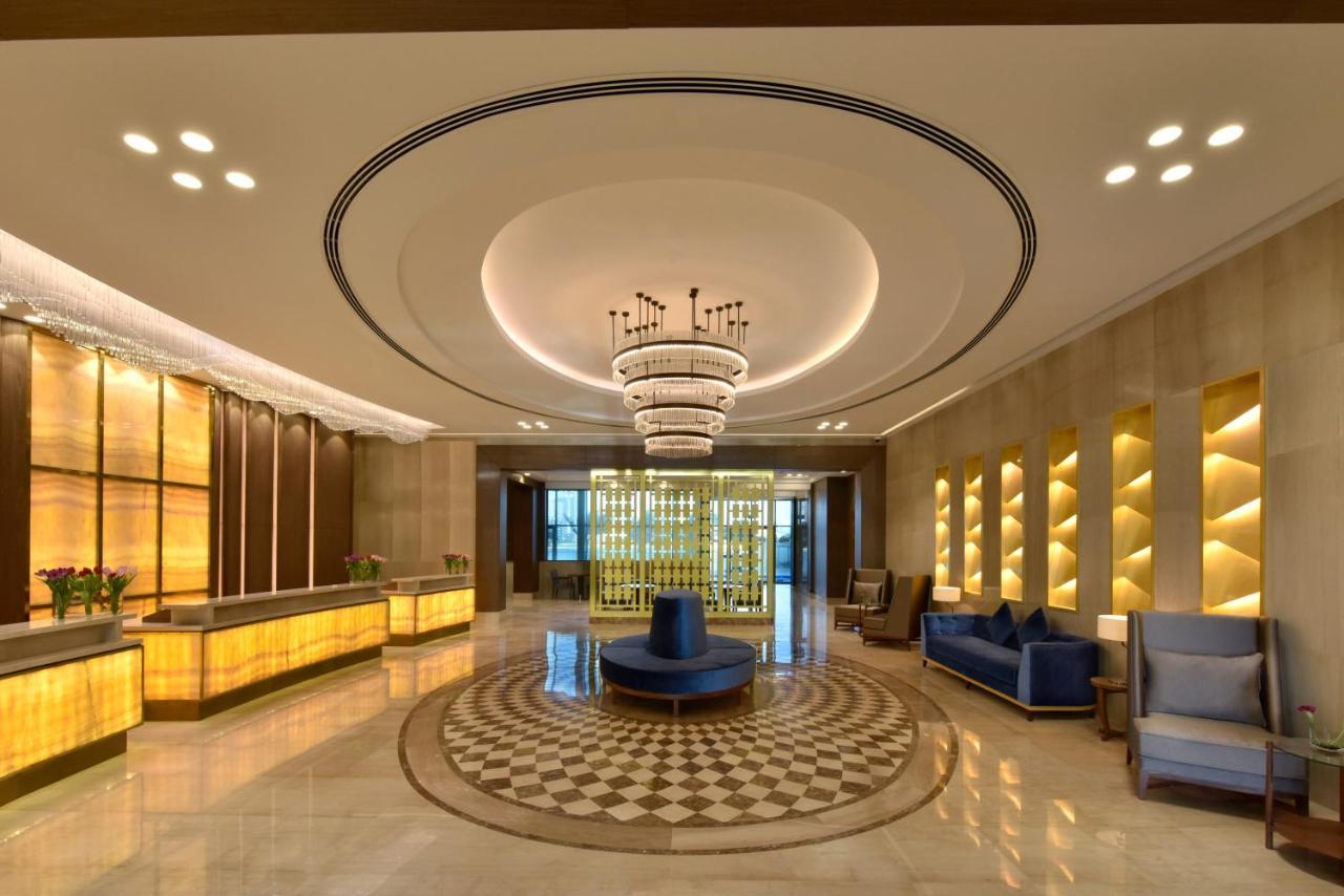 Grand Swiss-Belhotel Waterfront Seef Sanabis Exterior photo The image depicts a modern hotel lobby. It features a stylish design with warm lighting and elegant decor. There is a reception desk made of light-colored wood and illuminated panels. The ceiling has a large, circular light fixture, adding a luxuriou