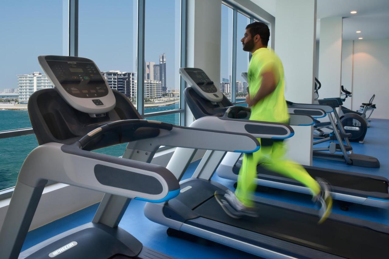Grand Swiss-Belhotel Waterfront Seef Sanabis Exterior photo The photo features a modern gym setting with several treadmills positioned next to large windows that showcase a cityscape and waterfront view. A man is running on one of the treadmills, wearing bright neon athletic attire. The overall atmosphere app