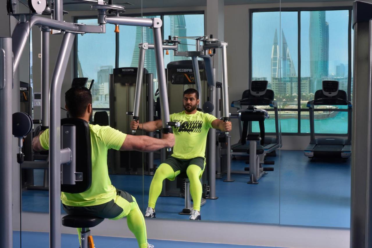 Grand Swiss-Belhotel Waterfront Seef Sanabis Exterior photo The photo shows a gym setting where a man is using a chest press machine. He is wearing bright yellow workout attire and is seated in front of a mirror that reflects his position. The gym has large windows that display a city skyline in the backgroun