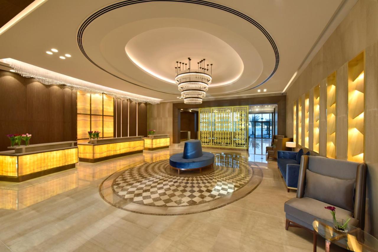 Grand Swiss-Belhotel Waterfront Seef Sanabis Exterior photo The image shows a modern hotel lobby. It features a spacious area with elegant design elements, including a large, decorative chandelier hanging from a curved ceiling. The reception desk is made of polished wood and illuminated by soft lighting. In t