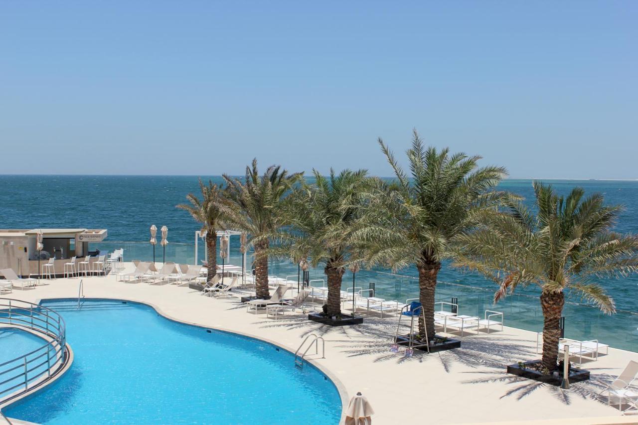 Grand Swiss-Belhotel Waterfront Seef Sanabis Exterior photo The photo depicts a scenic view of a tropical landscape with a swimming pool in the foreground. The pool has a curved shape and is surrounded by white lounge chairs. In the background, there are several tall palm trees providing shade. Beyond the poo