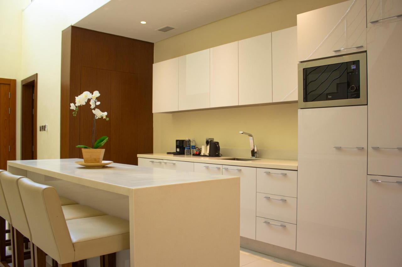 Grand Swiss-Belhotel Waterfront Seef Sanabis Exterior photo The photo shows a modern kitchen design featuring sleek white cabinets and countertops. On one side, there is a small island with bar stools, providing a space for seating. A potted plant, likely an orchid, is placed on the island. The kitchen is wel