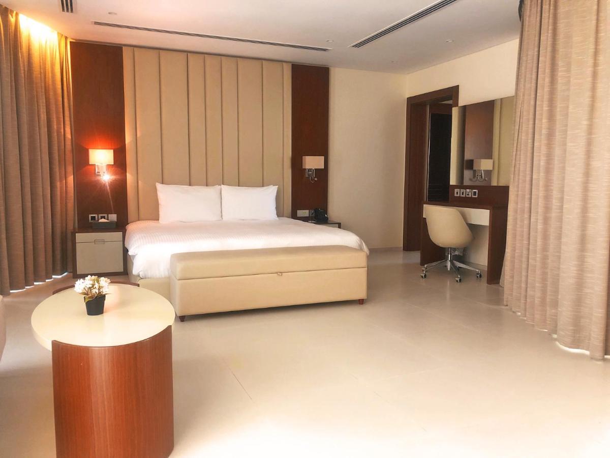 Grand Swiss-Belhotel Waterfront Seef Sanabis Exterior photo The photo shows a modern hotel room featuring a comfortable bed with white linens and pillows placed against a backdrop of muted, vertical wall paneling. The room is well-lit, with warm lighting coming from wall sconces and large windows covered by f