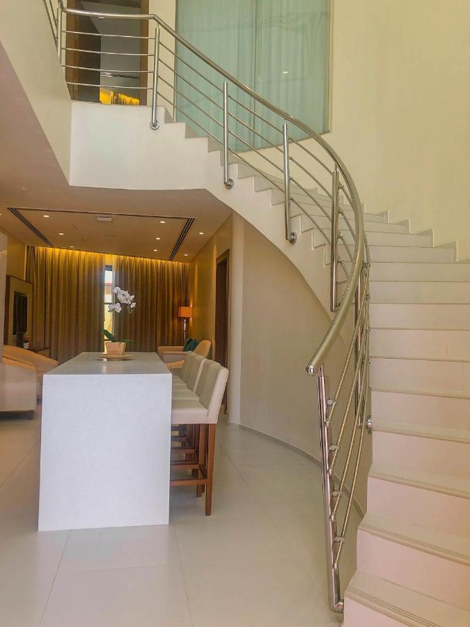 Grand Swiss-Belhotel Waterfront Seef Sanabis Exterior photo The photo shows a modern interior space with an open design. On the left, there is a sleek white bar-style counter with several stools arranged around it. To the right, there is a gracefully curved staircase leading to an upper level, featuring a met