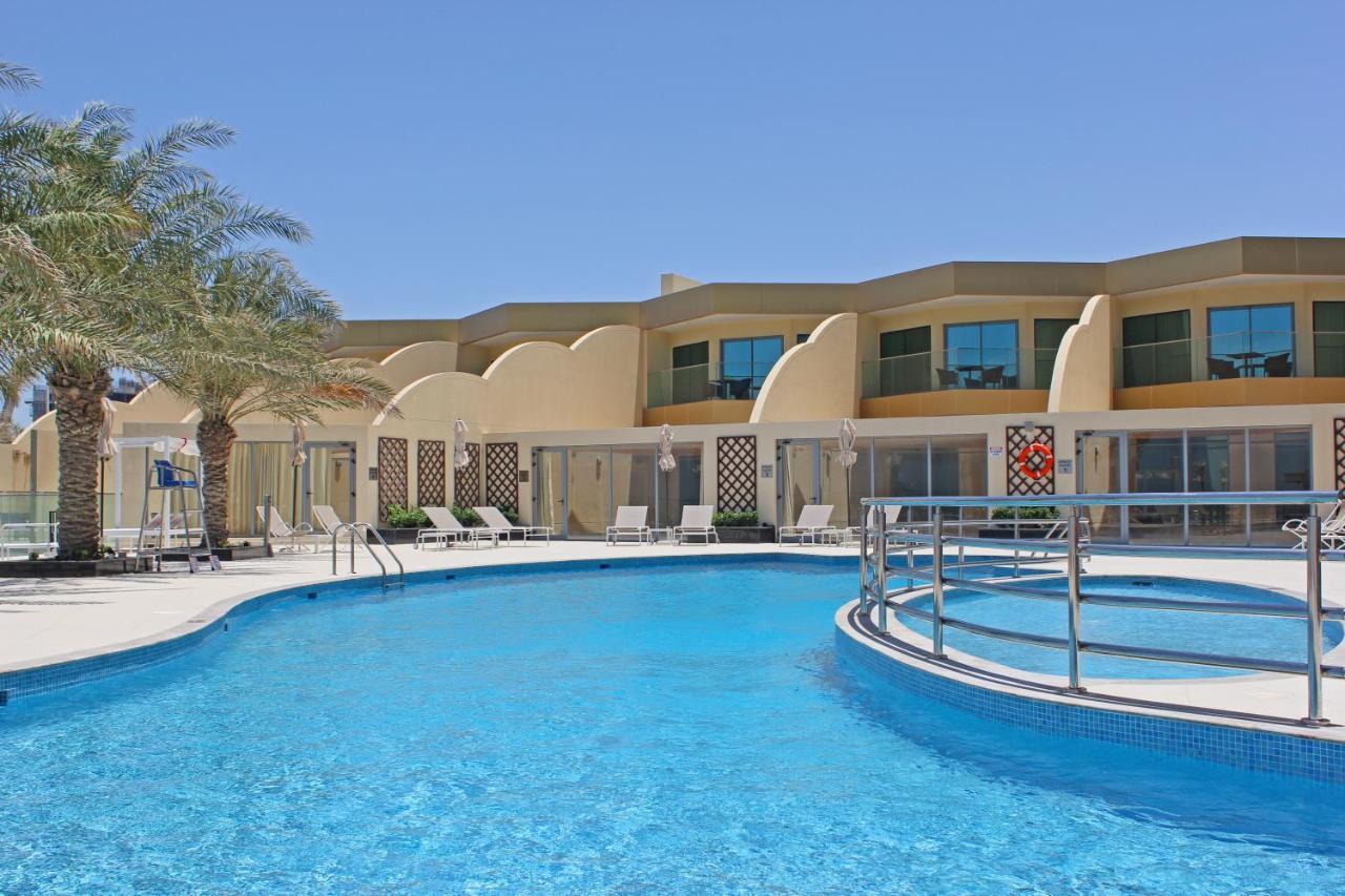 Grand Swiss-Belhotel Waterfront Seef Sanabis Exterior photo The photo shows a swimming pool with clear blue water, surrounded by a deck. There are lounge chairs arranged around the pool. In the background, there is a building with a modern architectural design featuring large windows and a light-colored facad