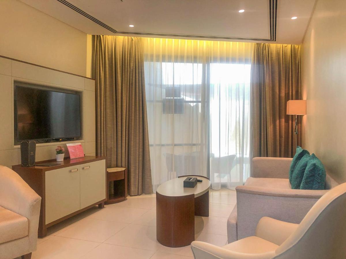 Grand Swiss-Belhotel Waterfront Seef Sanabis Exterior photo The photo shows a modern hotel room or a living space. It features a comfortable seating area with two white chairs and a small, round wooden coffee table. There is a flat-screen television mounted on the wall, with a console or cabinet below it. Lig
