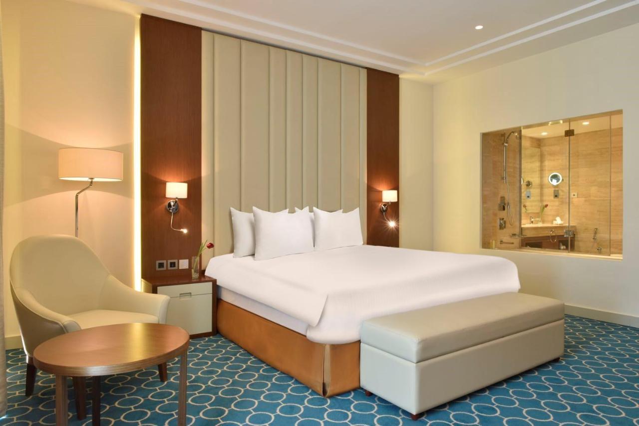 Grand Swiss-Belhotel Waterfront Seef Sanabis Exterior photo The photo shows a modern hotel room. It features a large bed with white bedding, placed against a wall with a textured design. To the side, there is a comfortable chair and a small round table. There is also a bench at the foot of the bed. The room h