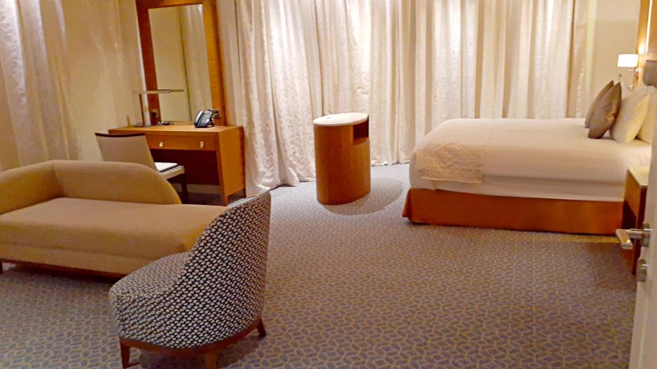 Grand Swiss-Belhotel Waterfront Seef Sanabis Exterior photo The photo shows a modern hotel room with a minimalistic design. There is a large bed against one wall with a simple headboard. Next to the bed, there is a small round table. On the opposite side, there is a comfortable-looking chair with a patterned 