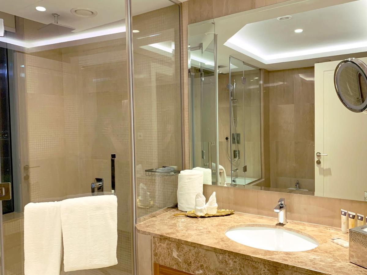 Grand Swiss-Belhotel Waterfront Seef Sanabis Exterior photo The photo shows a modern bathroom with a sleek design. There is a large mirror extending across the wall, reflecting the light fixtures above. The shower area is enclosed in glass, indicating a spacious shower. A marble countertop features a circular