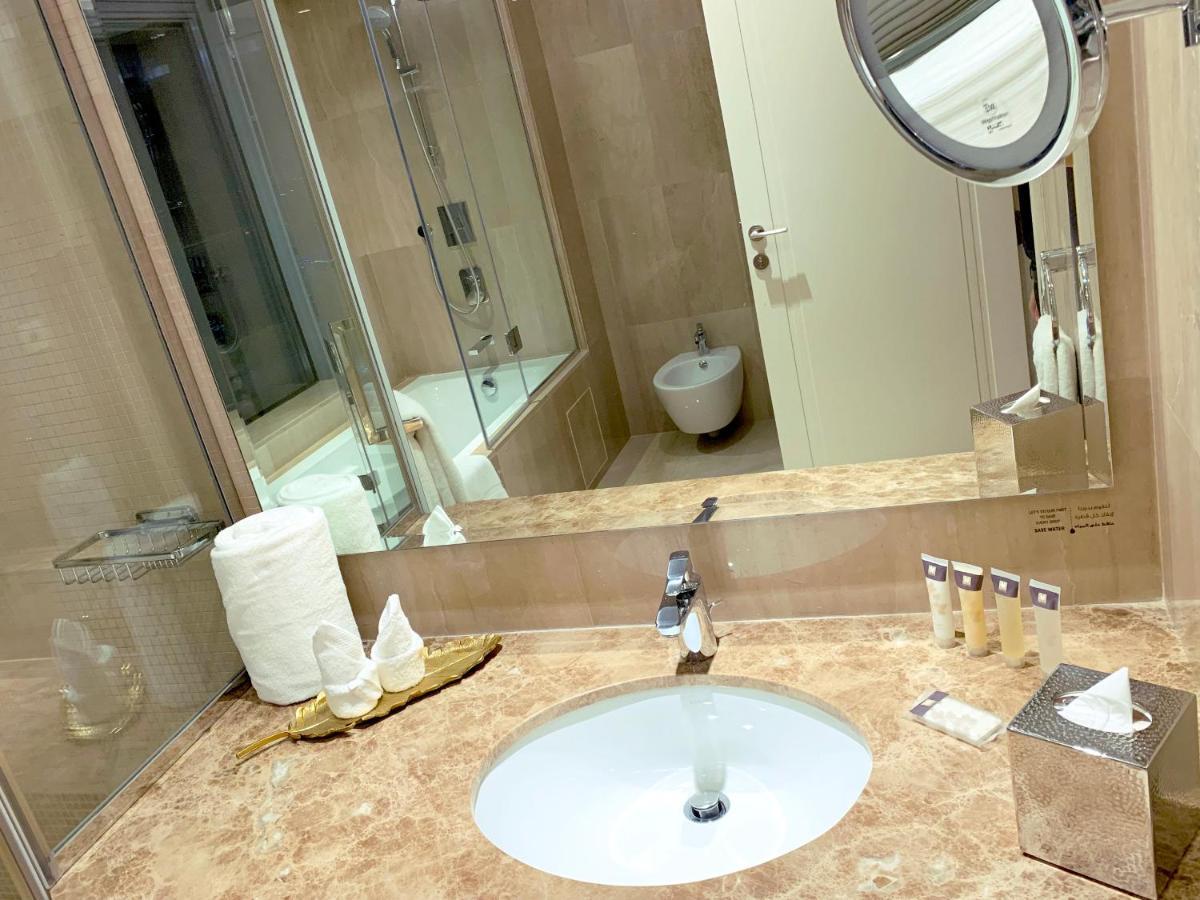 Grand Swiss-Belhotel Waterfront Seef Sanabis Exterior photo The photo shows a modern bathroom featuring a marble countertop with a circular sink and a faucet. There are neatly arranged toiletries on the countertop, including small bottles and disposable cups. A roll of toilet paper is visible, as well as a sm