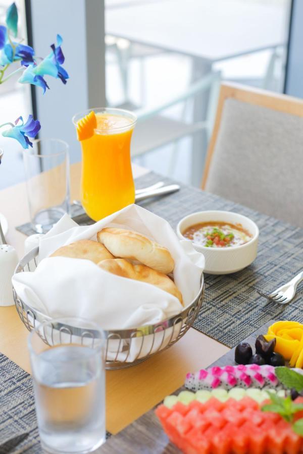 Grand Swiss-Belhotel Waterfront Seef Sanabis Exterior photo The photo shows a dining setup with a variety of food and drink. In the foreground, there is a basket lined with a white cloth containing several pieces of bread. Next to it, there’s a small bowl of a dish that appears to be a sauce or stew, garnishe