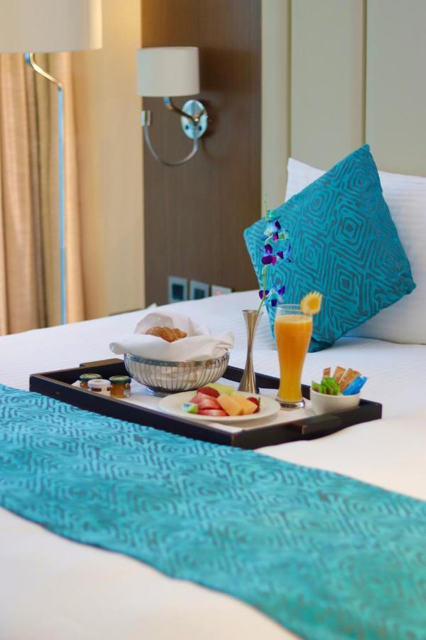 Grand Swiss-Belhotel Waterfront Seef Sanabis Exterior photo The photo shows a tray placed on a bed. On the tray, there is a variety of food items, including a basket with a pastry, sliced fruits, and small jars, likely containing condiments or spreads. There is also a tall glass filled with a bright orange be