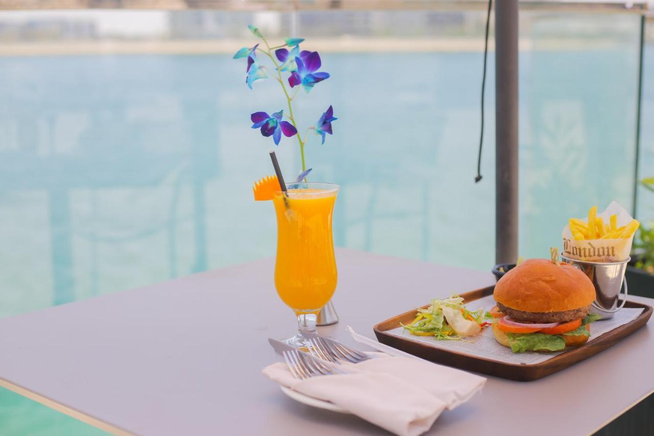 Grand Swiss-Belhotel Waterfront Seef Sanabis Exterior photo The photo features a refreshing scene with a vibrant orange drink garnished with a slice of orange and purple flowers, served in a glass. Next to it, there is a tray containing a hamburger with lettuce and possibly other condiments, accompanied by a 