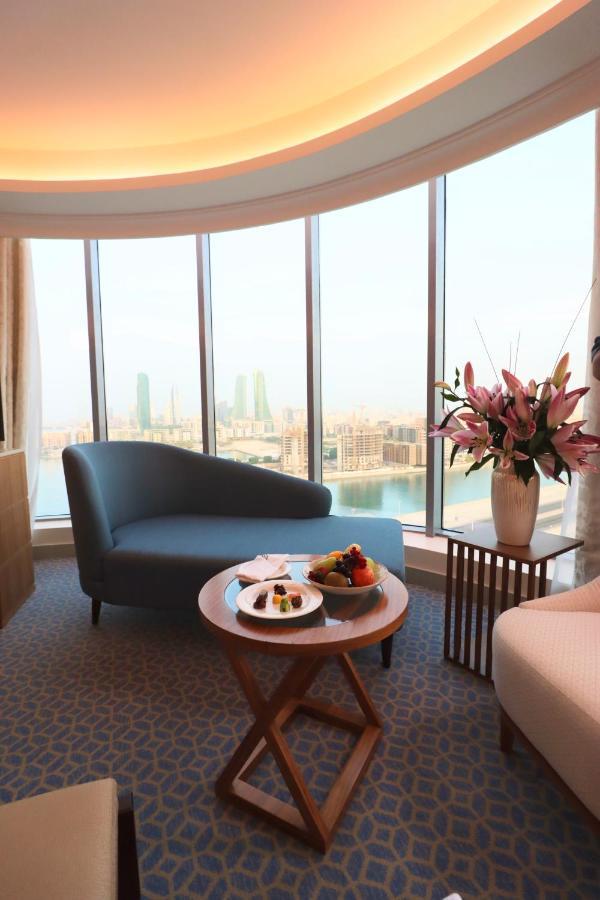 Grand Swiss-Belhotel Waterfront Seef Sanabis Exterior photo The image shows a stylish and modern interior of a hotel room or lounge area. There is a curved blue sofa positioned near large windows that offer a view of a cityscape, featuring tall buildings and what appears to be water in the foreground. A round