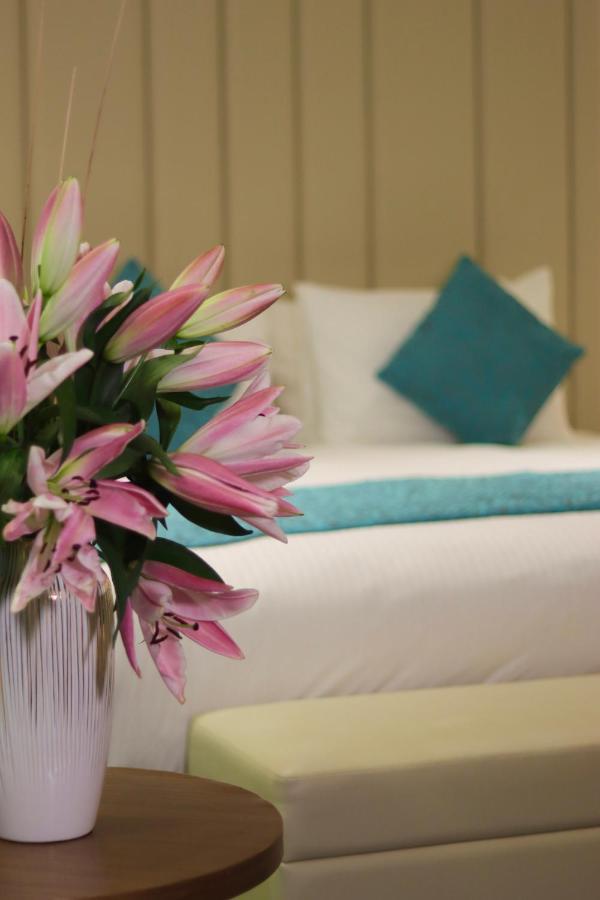 Grand Swiss-Belhotel Waterfront Seef Sanabis Exterior photo The image features a close-up of a bouquet of pink lilies positioned in the foreground, creating a vibrant and fresh atmosphere. In the background, there is a neatly made bed with white linens and a decorative turquoise pillow, adding a touch of colo