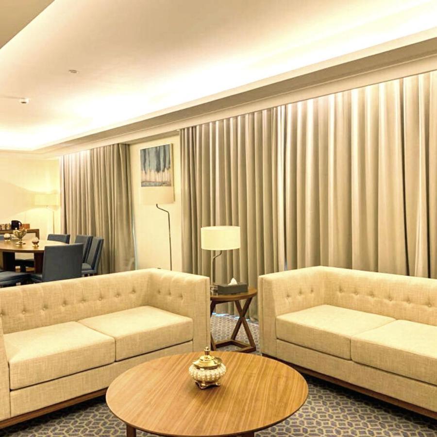 Grand Swiss-Belhotel Waterfront Seef Sanabis Exterior photo The photo shows a modern, softly lit living area featuring two light-colored sofas with simple, elegant designs. In the center, there is a round wooden coffee table adorned with a decorative item. The walls are adorned with large, neutral-toned curta