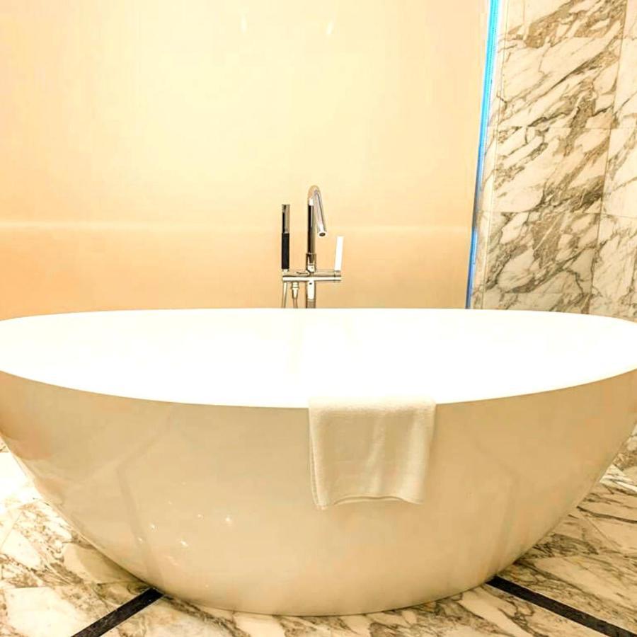 Grand Swiss-Belhotel Waterfront Seef Sanabis Exterior photo The photo shows a modern, oval-shaped bathtub located in a bathroom. The tub is white and has a sleek, minimalist design. A chrome faucet with a handle is mounted above the tub. There’s a white towel draped over the edge of the tub. The surrounding w