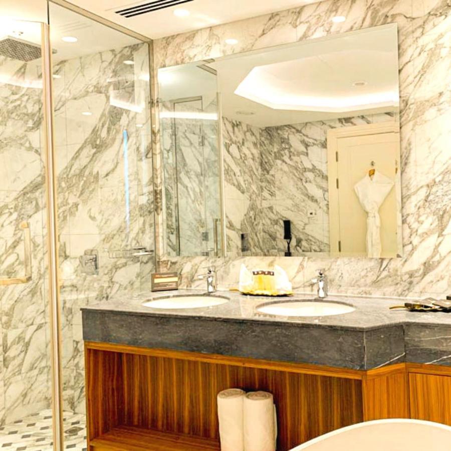 Grand Swiss-Belhotel Waterfront Seef Sanabis Exterior photo The photo depicts a luxurious bathroom featuring elegant marble walls. There are two sinks with a dark granite countertop, and the cabinetry below has a warm wooden finish. Large mirrors are mounted above the sinks, reflecting the stylish design. A w